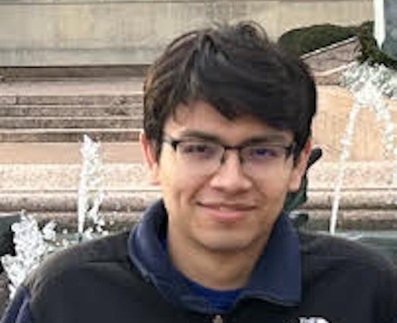 man with dark hair and glasses smiling by a fountain