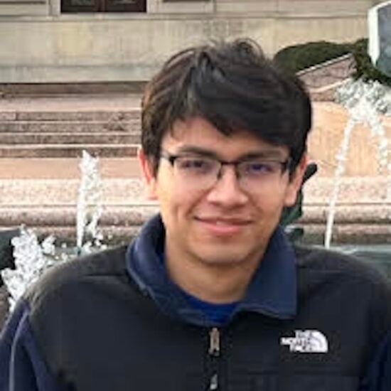 man with dark hair and glass smiling by a fountain